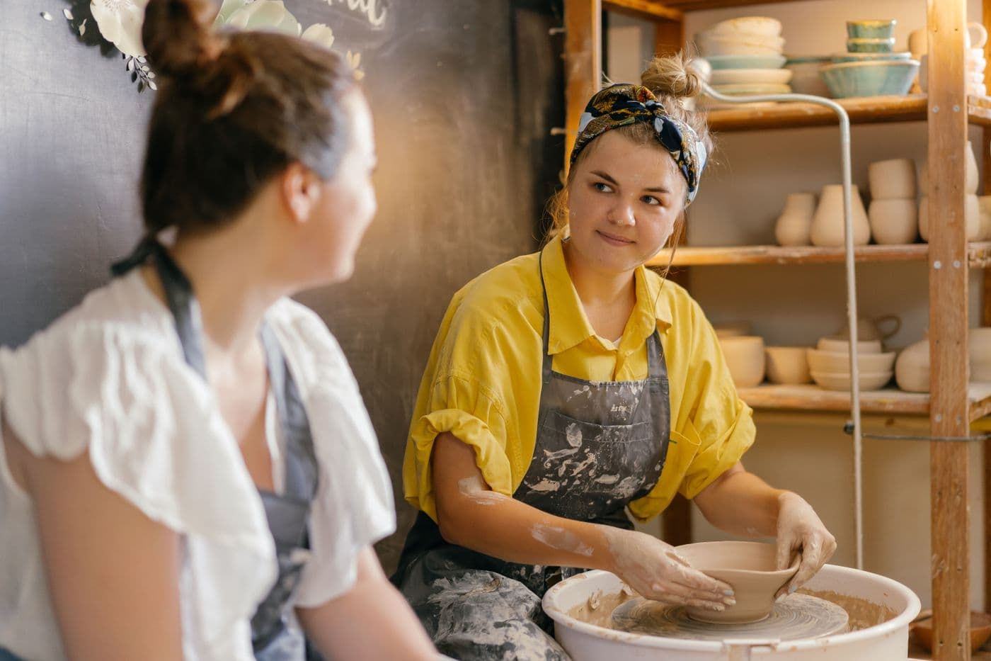 Two women working together at a pottery wheel, surrounded by handmade ceramics
